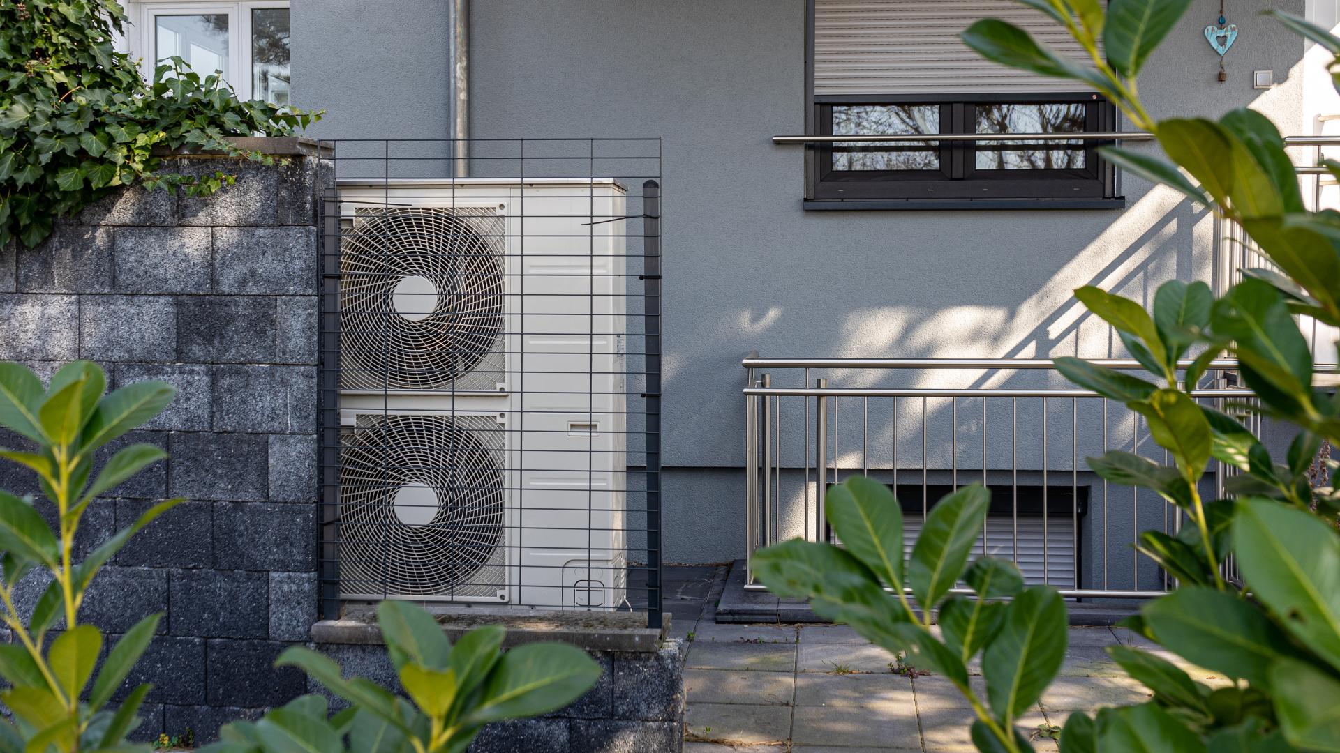 Unidad exterior de bomba de calor de aerotermia instalada junto a una vivienda moderna con jardín. Sistema de aerotermia eficiente para climatización de viviendas.